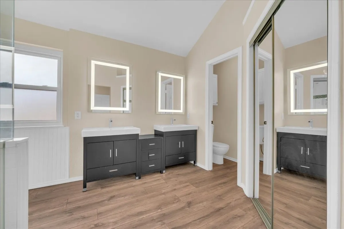 Dual vanities with integrated sinks, illuminated mirrors, wood-finish flooring, and a mirrored sliding closet door