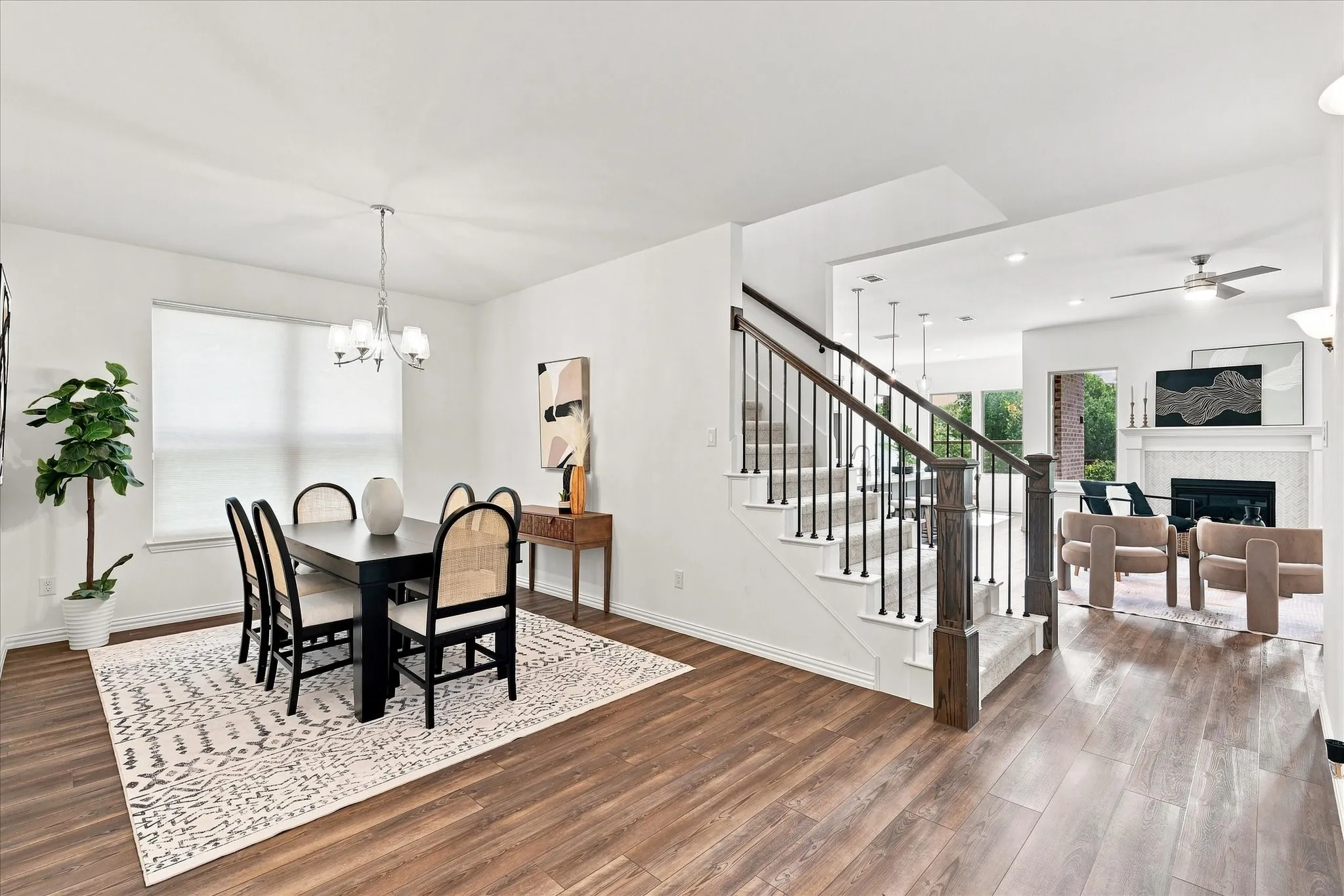 Wood-finish flooring throughout, a staircase with dark wood newel posts and black iron balusters, a built-in fireplace with a mantel, and a dining area with a chandelier