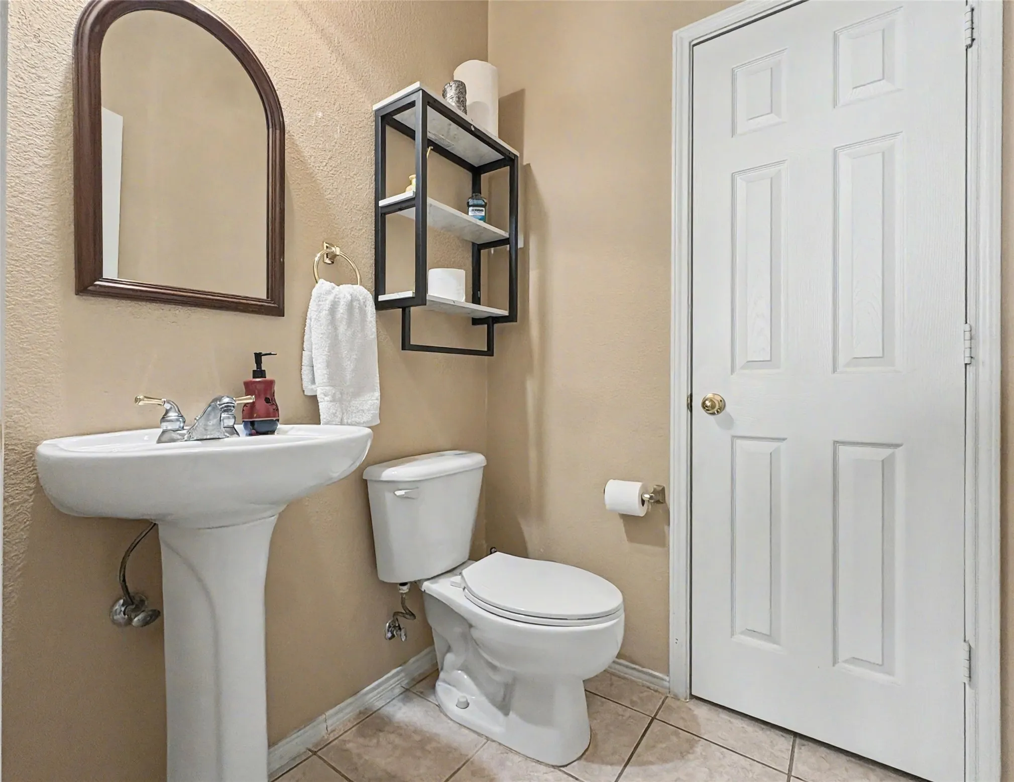 Pedestal sink with polished chrome faucet, arched framed mirror, wall-mounted shelving unit, and tile flooring