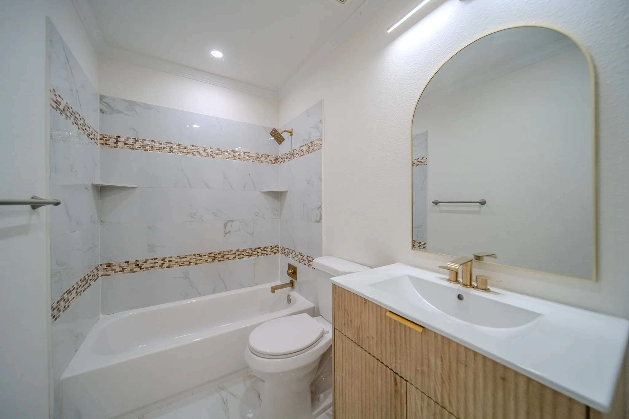 Recessed lighting, wood-finish vanity with gold-tone hardware, rectangular undermount sink, arched mirror, and white tiled shower with decorative mosaic bands and built-in shelves