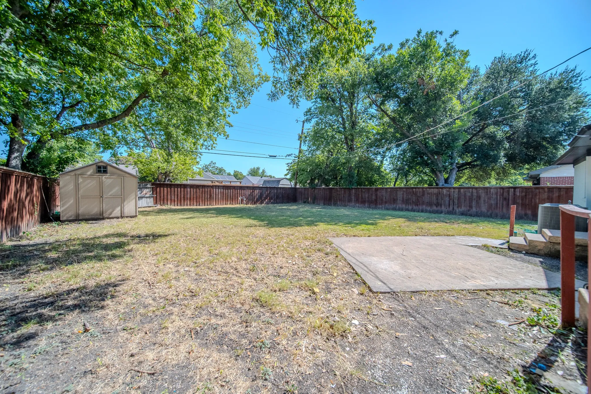 Expansive backyard featuring a privacy fence, mature trees, concrete patio, and storage shed