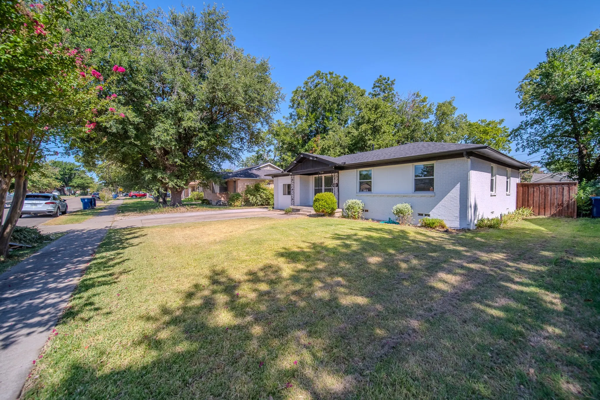 White brick exterior, dark roof, established tree canopy, green lawn, and privacy fencing