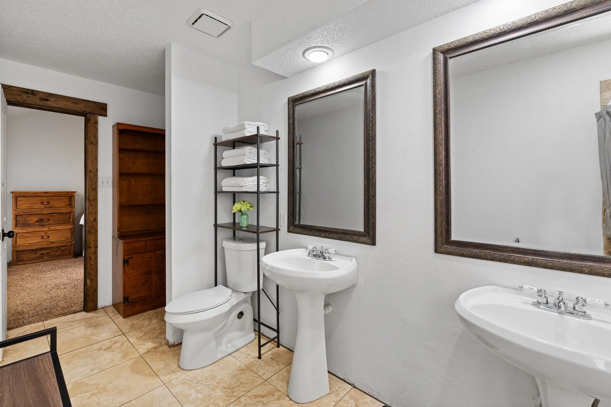 Dual pedestal sinks, framed mirrors, and a built-in wood-finish shelving unit complement the tile flooring