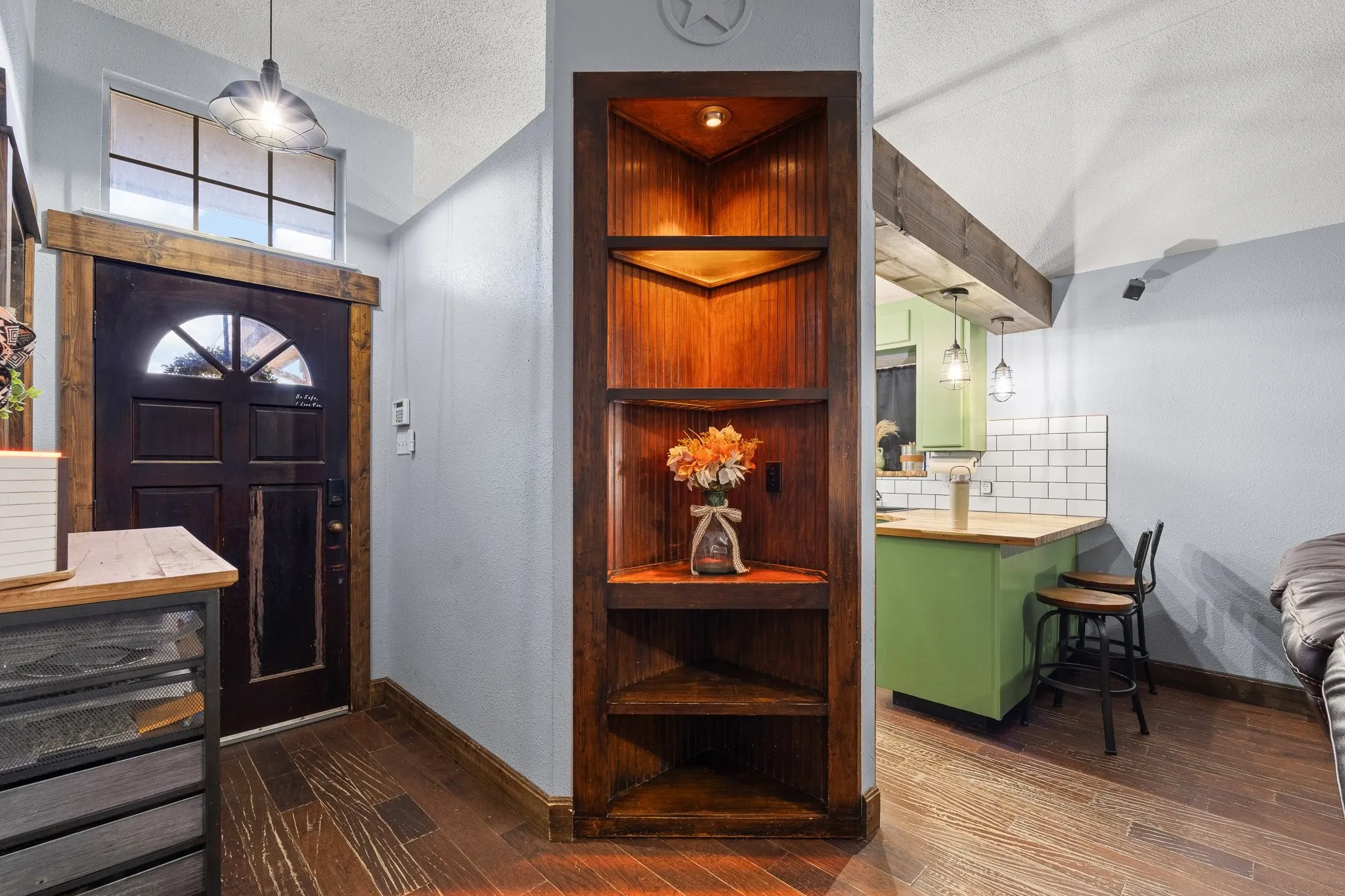 Wood-finish flooring throughout, dark wood-paneled corner shelving, a dark wood entry door with a decorative arch, and a kitchen area with green cabinetry and subway tile backsplash