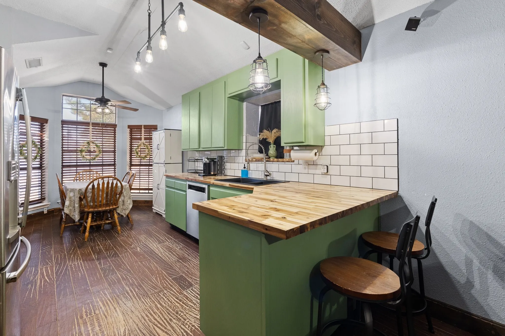 Vaulted ceilings with exposed wood beams, wood-finish flooring, custom green cabinetry, white subway tile backsplash, and light wood countertops