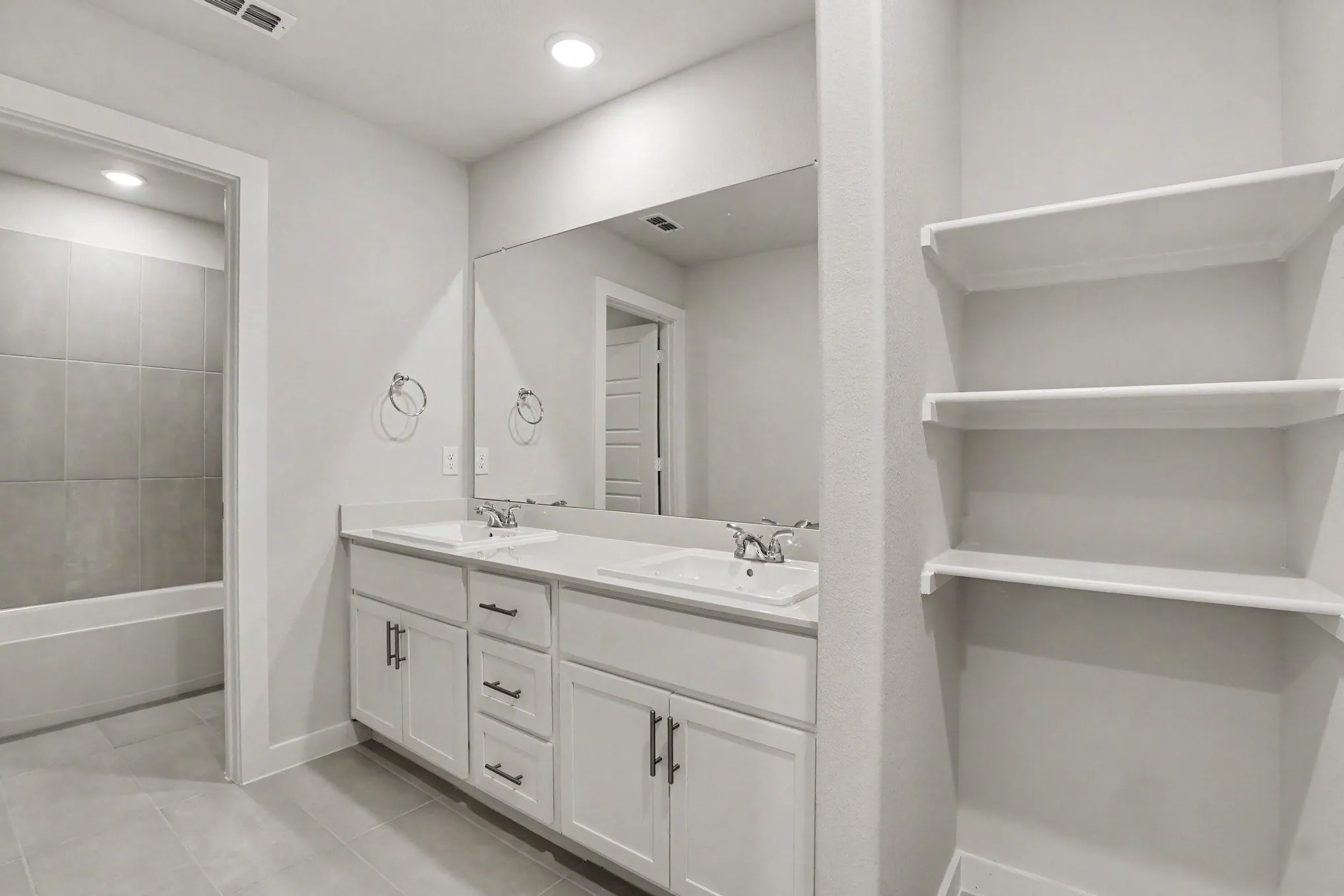 Dual sink vanity with white cabinetry and dark hardware, large vanity mirror, built-in shelving, and tiled flooring