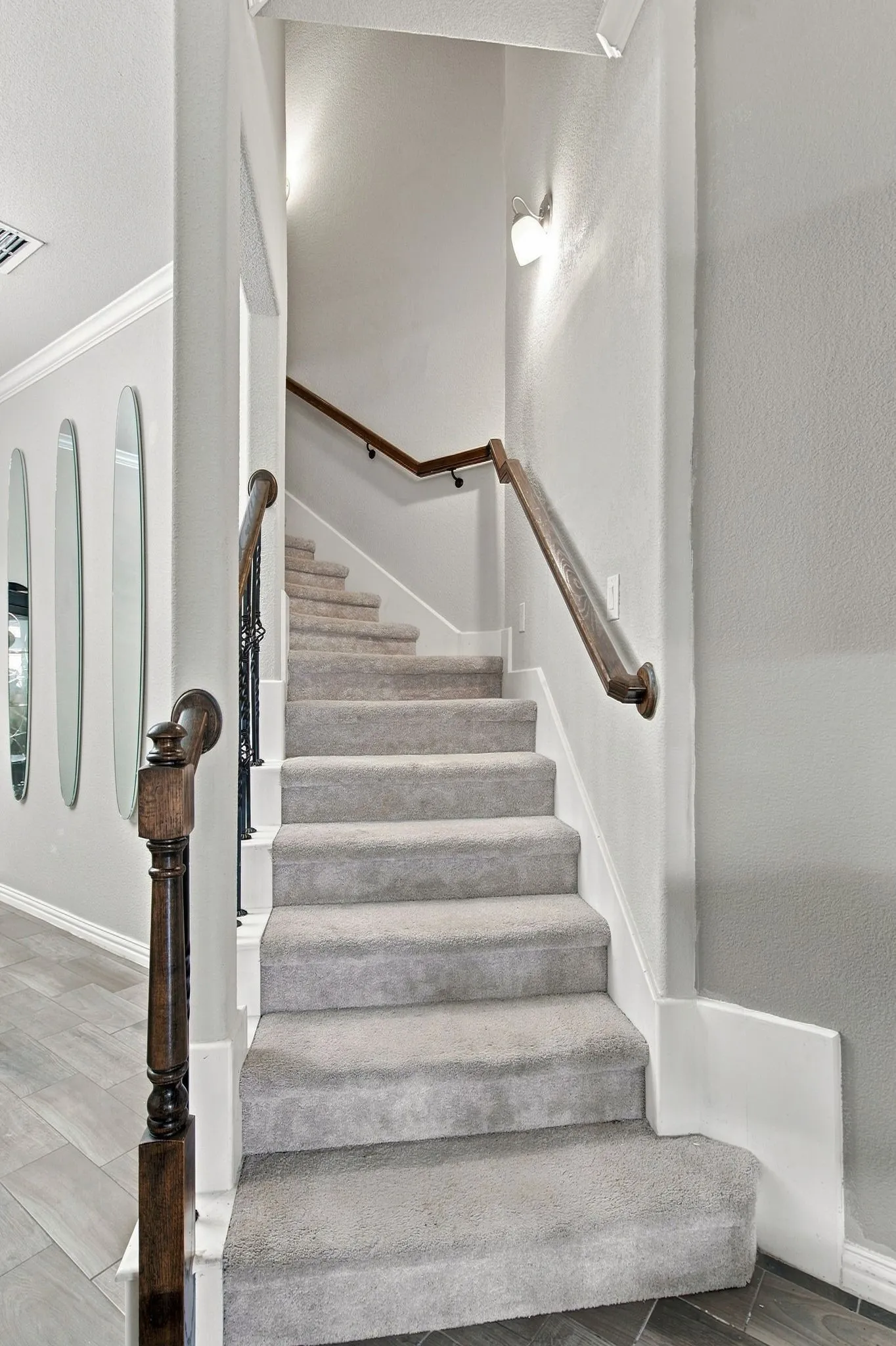 Carpeted staircase featuring a dark wood handrail and balusters, with a wall-mounted light fixture