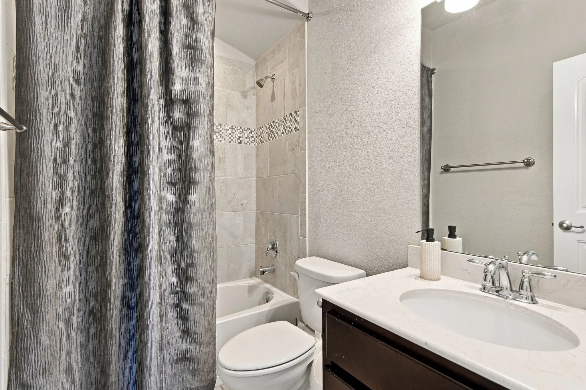 Tiled tub-shower combination with decorative accent band, dark wood-finish vanity, undermount sink, and polished chrome fixtures
