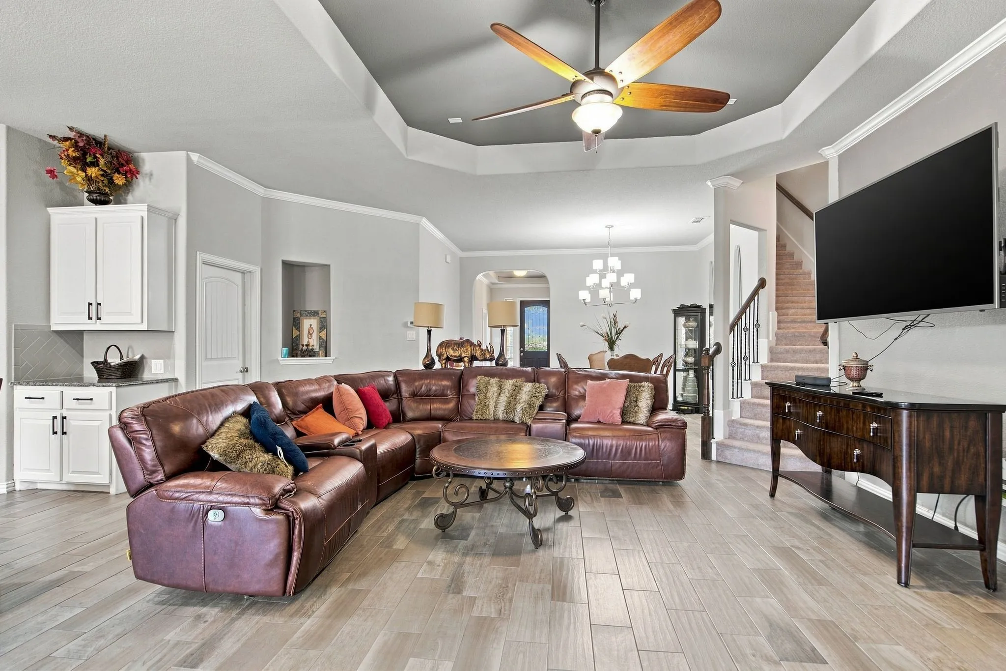Tray ceiling with a ceiling fan, wood-finish tile flooring, built-in display niche, white cabinetry with dark hardware, and a staircase with dark wood handrails