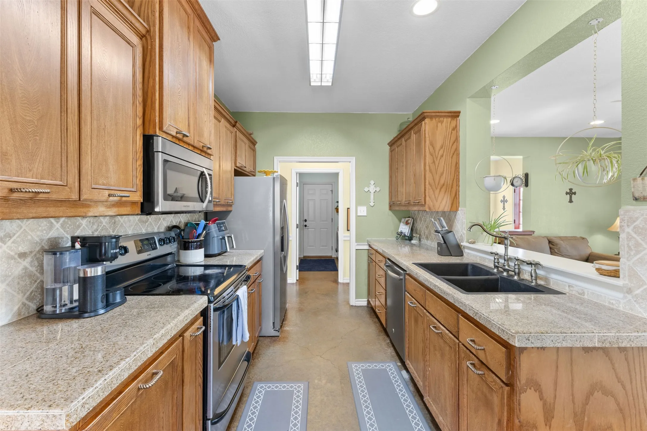 Galley kitchen featuring wood cabinetry, stone-finish countertops, stainless steel appliances, a tile backsplash, and a pass-through opening