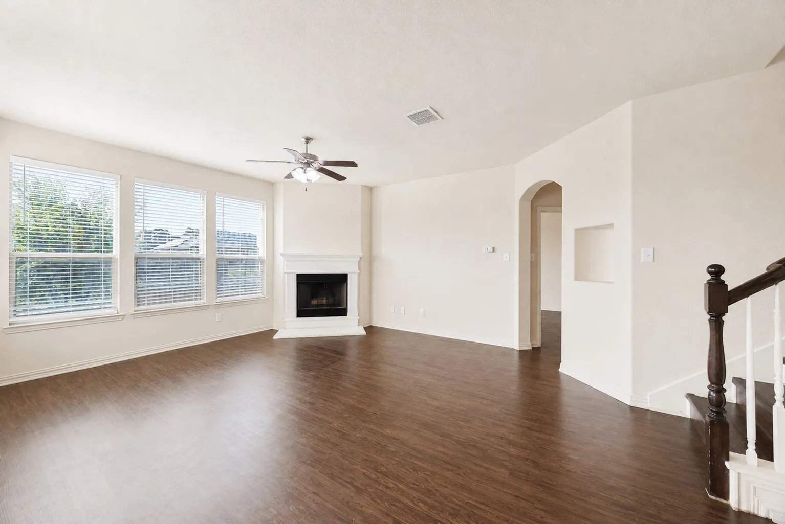 Wood-finish flooring, multiple windows with blinds, built-in fireplace, ceiling fan, and staircase with wood newel post and handrail