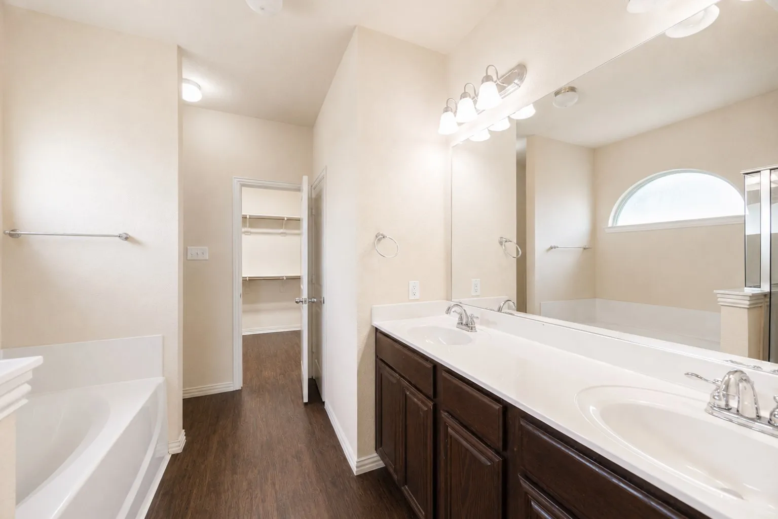 Dual vanity with integrated sinks, wood-finish cabinetry, a large mirror, a built-in bathtub, and an arched window