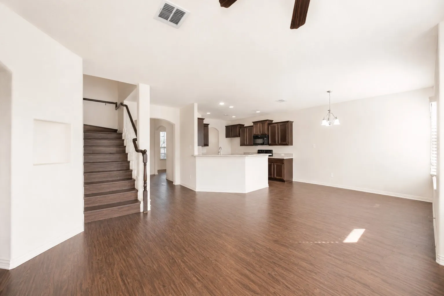 Open-concept living area featuring wood-finish flooring, a kitchen with dark wood cabinetry and white countertops, a staircase with dark wood treads and a white banister, and a ceiling fan