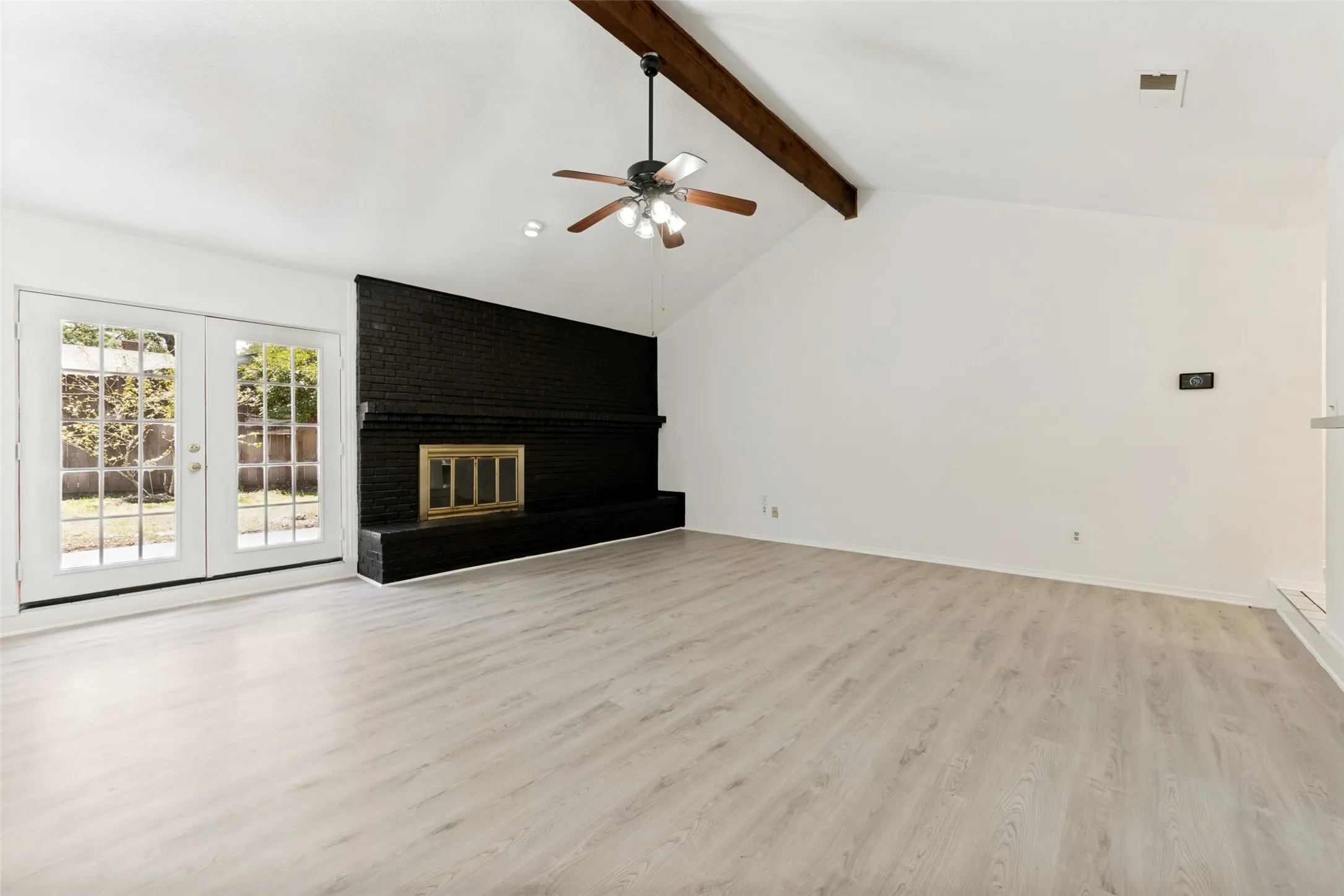 Vaulted ceiling with exposed wood beam, black brick fireplace with hearth, wood-finish flooring, and French doors