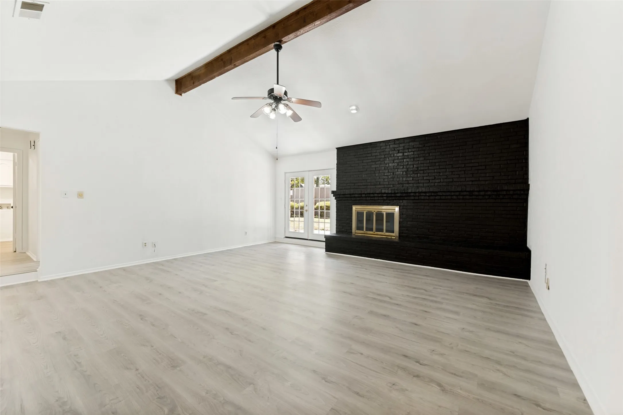 Vaulted ceiling with exposed wood beam, wood-finish flooring, black brick fireplace with a brass firebox, and French doors