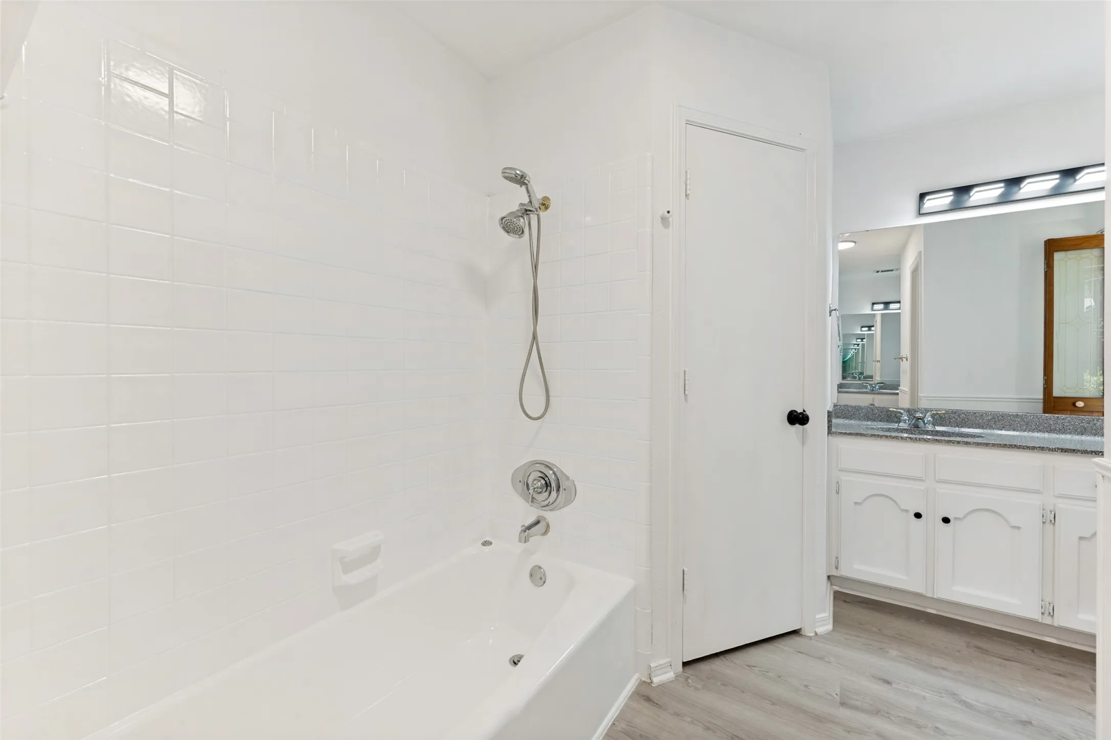 White tile shower surround with chrome fixtures, a built-in bathtub, light wood-finish flooring, a white vanity with a light-colored countertop, and a vanity mirror with overhead lighting