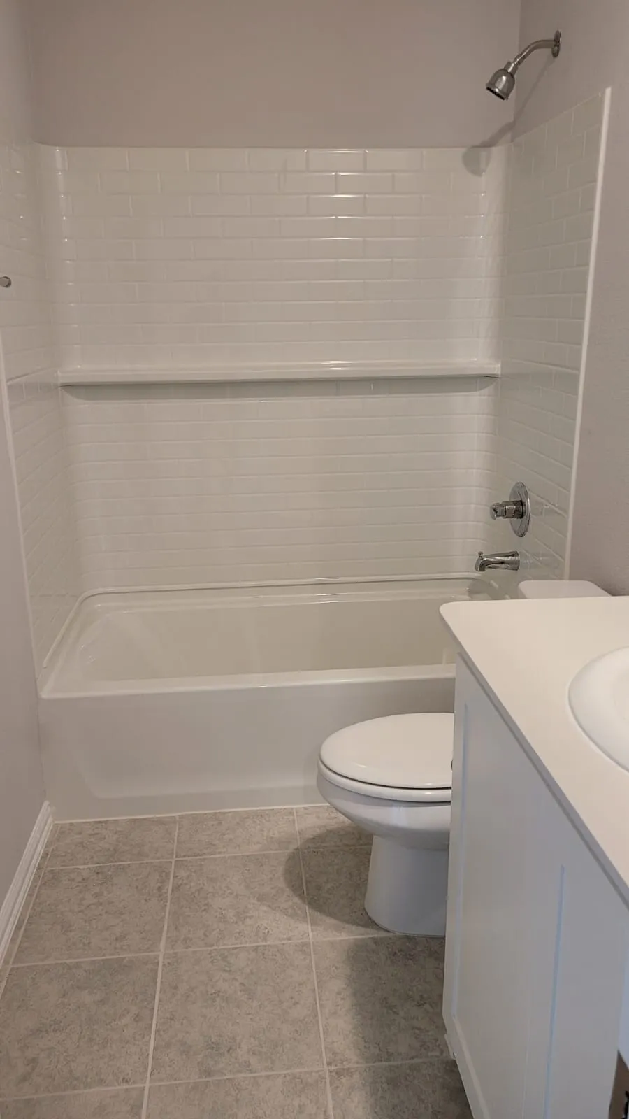 White subway tile shower surround with integrated shelf, white bathtub, chrome shower fixtures, tile flooring, and white vanity with integrated sink