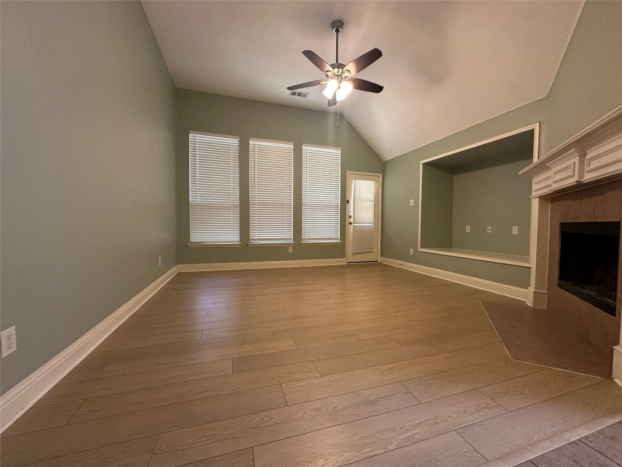 Vaulted ceiling with ceiling fan, three paneled windows, wood-finish flooring, built-in entertainment niche, and a fireplace with decorative mantle