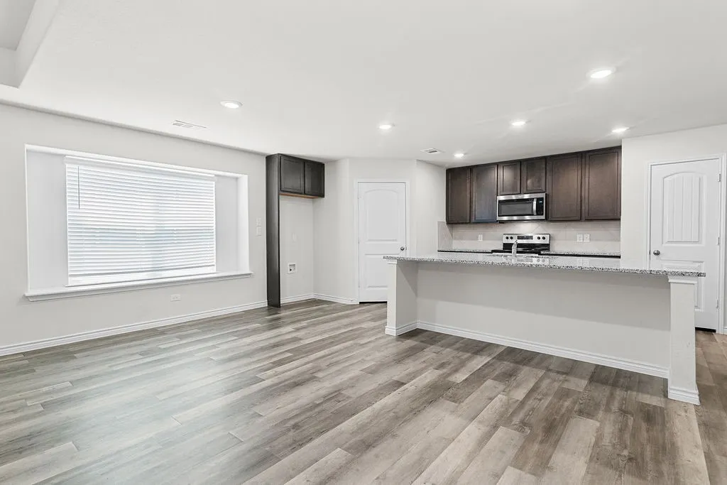 Spacious open-concept area featuring wood-finish flooring, recessed lighting, a kitchen island with a light-colored countertop, dark wood cabinetry, and stainless steel appliances