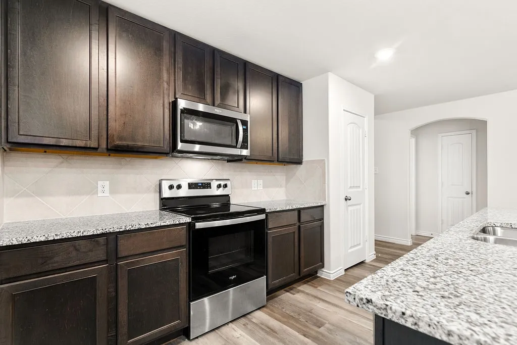 Dark wood-finish cabinetry, stainless steel appliances, speckled stone countertops, tile backsplash, and wood-finish flooring