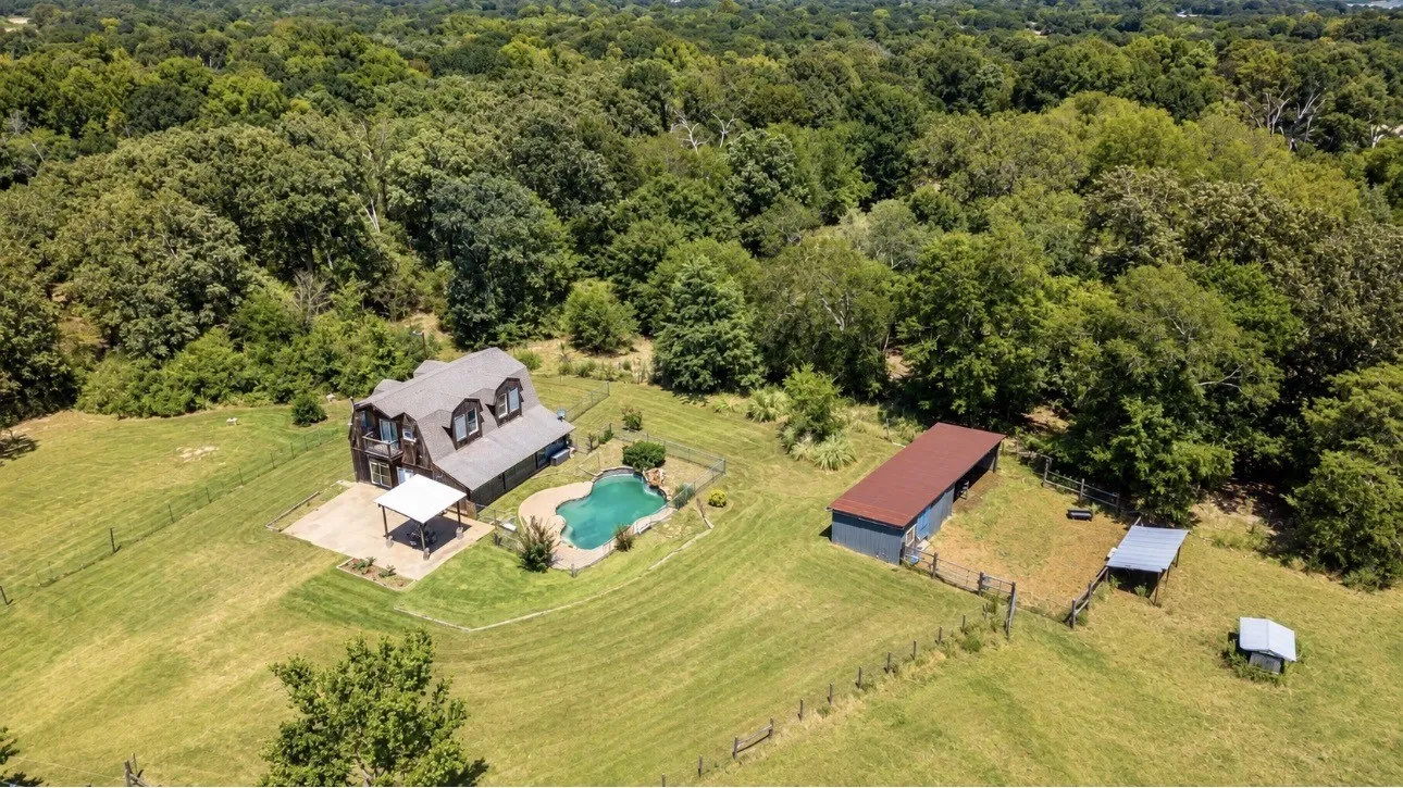 Gabled roofline residence with a swimming pool, covered patio, barn, and fenced pasture surrounded by mature trees
