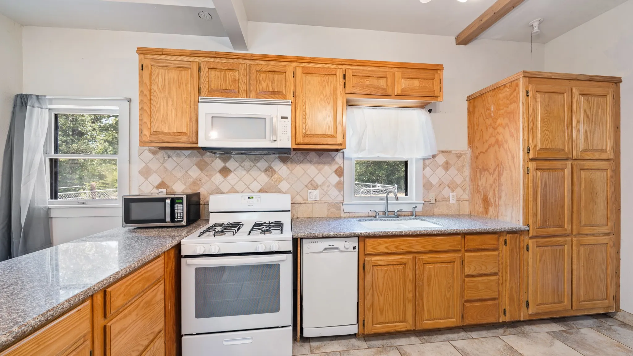 Kitchen featuring wood cabinetry, light-toned countertops, a tile backsplash, and white appliances