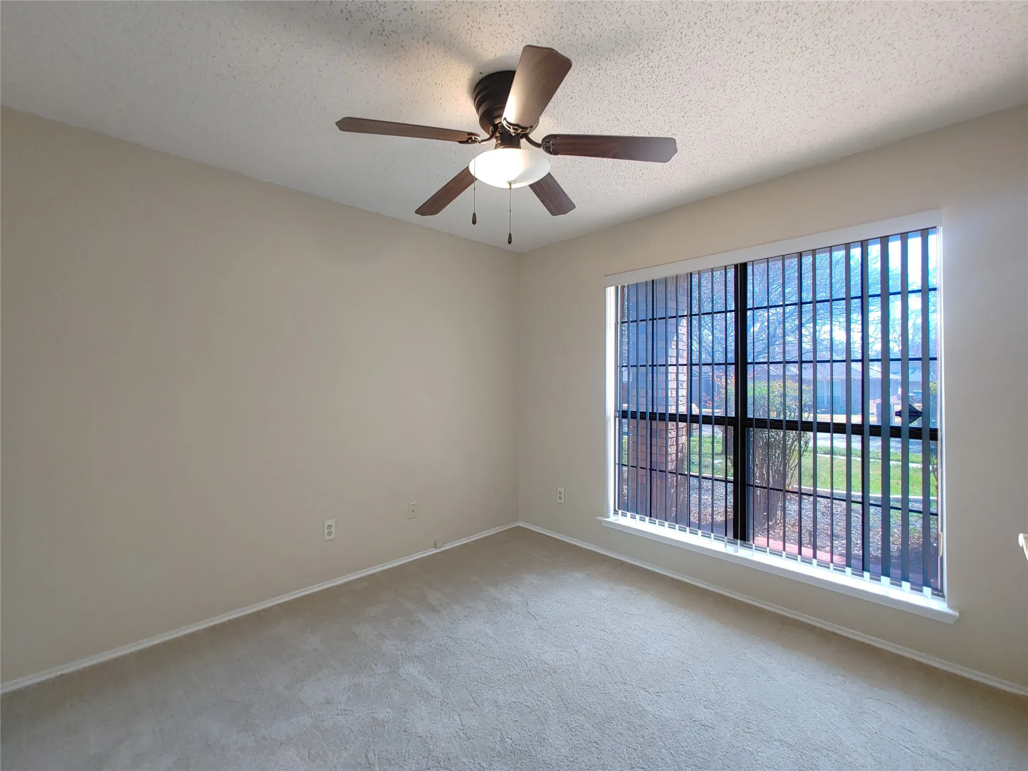 Textured ceiling with a ceiling fan light fixture, beige carpet, and a large window with vertical blinds