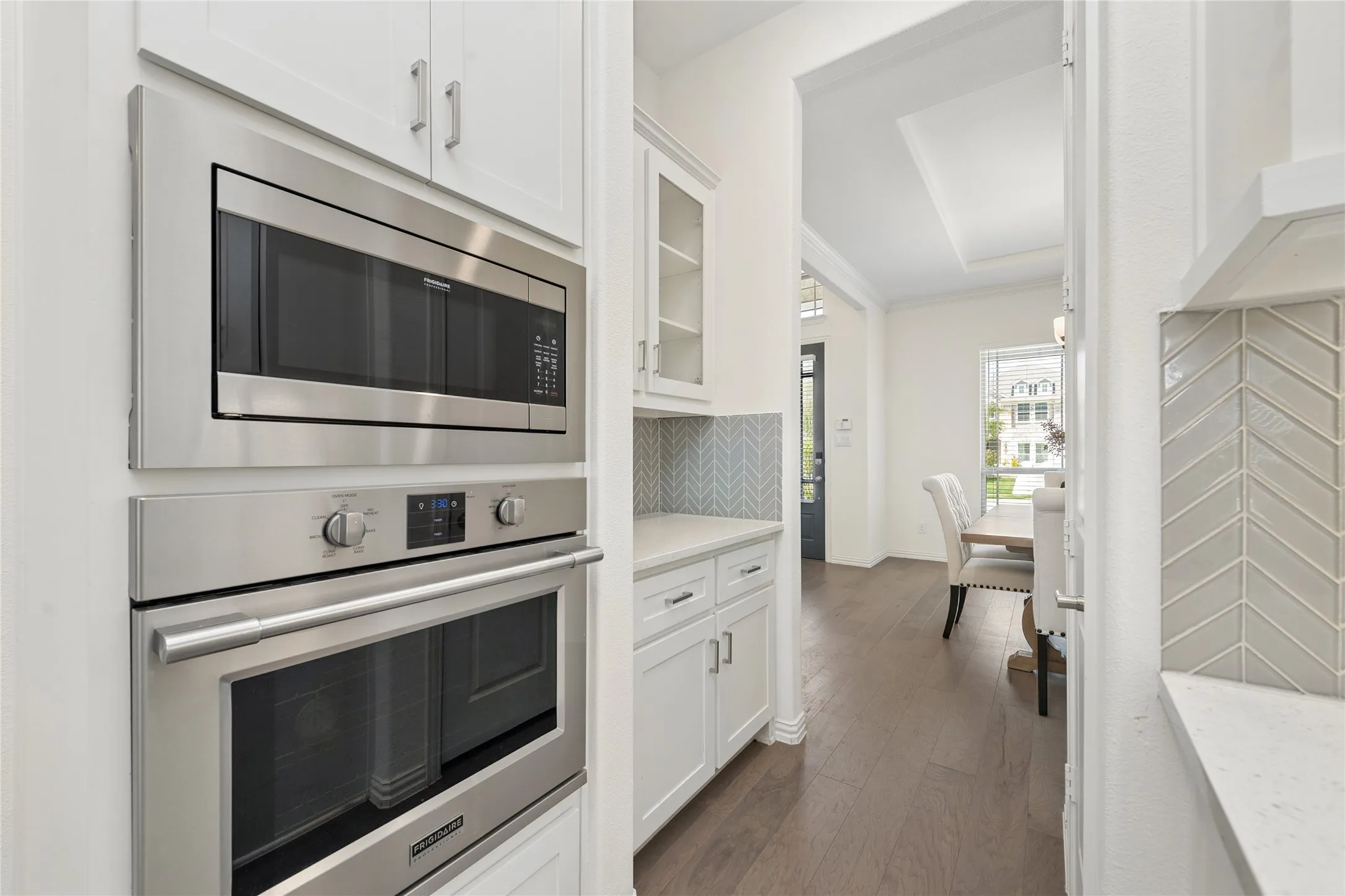 Built-in stainless steel microwave and oven, white cabinetry, chevron tile backsplash, wood-finish flooring, and tray ceiling detail