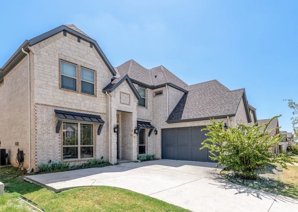 Stone and brick facade, dark roofing, attached garage, concrete driveway, and black window awnings