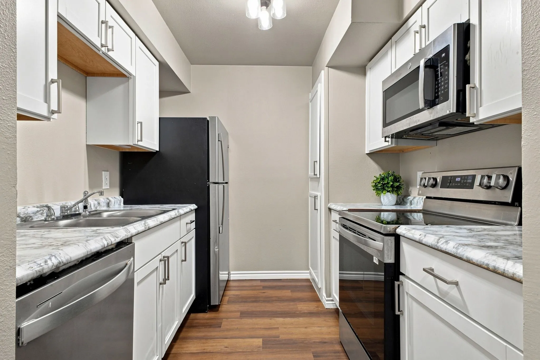 Galley kitchen featuring white cabinetry with chrome hardware, marble-finish countertops, stainless steel appliances including a gas range and microwave, black refrigerator, and wood-finish flooring throughout