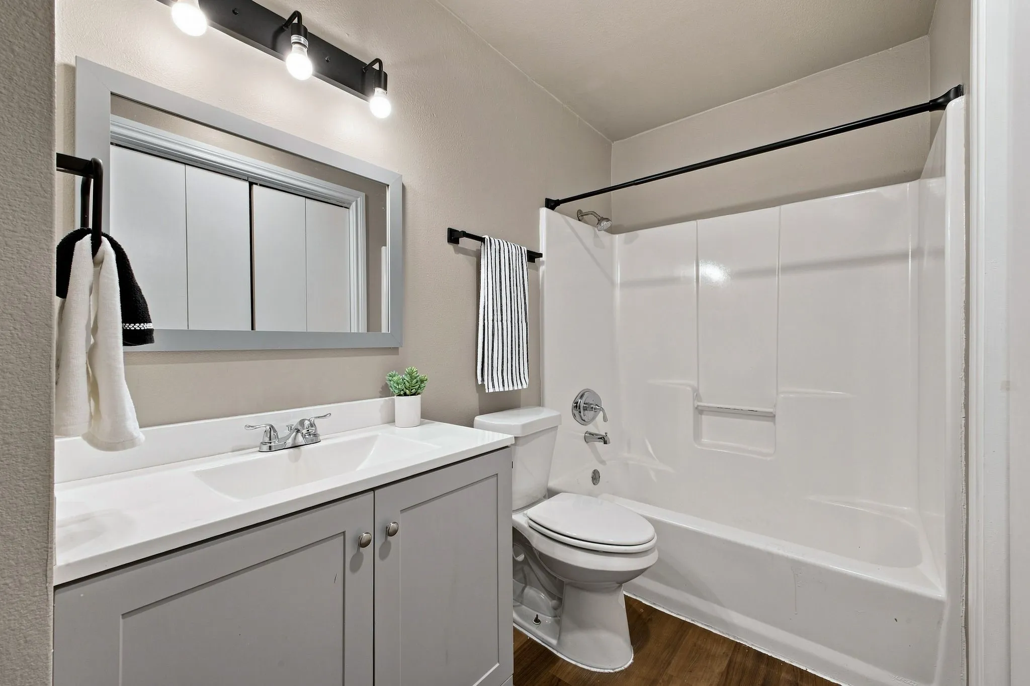 Bathroom featuring wood-finish flooring, a white integrated sink with chrome fixtures, a gray vanity with cabinet storage, a framed mirror, and a white tub/shower combination