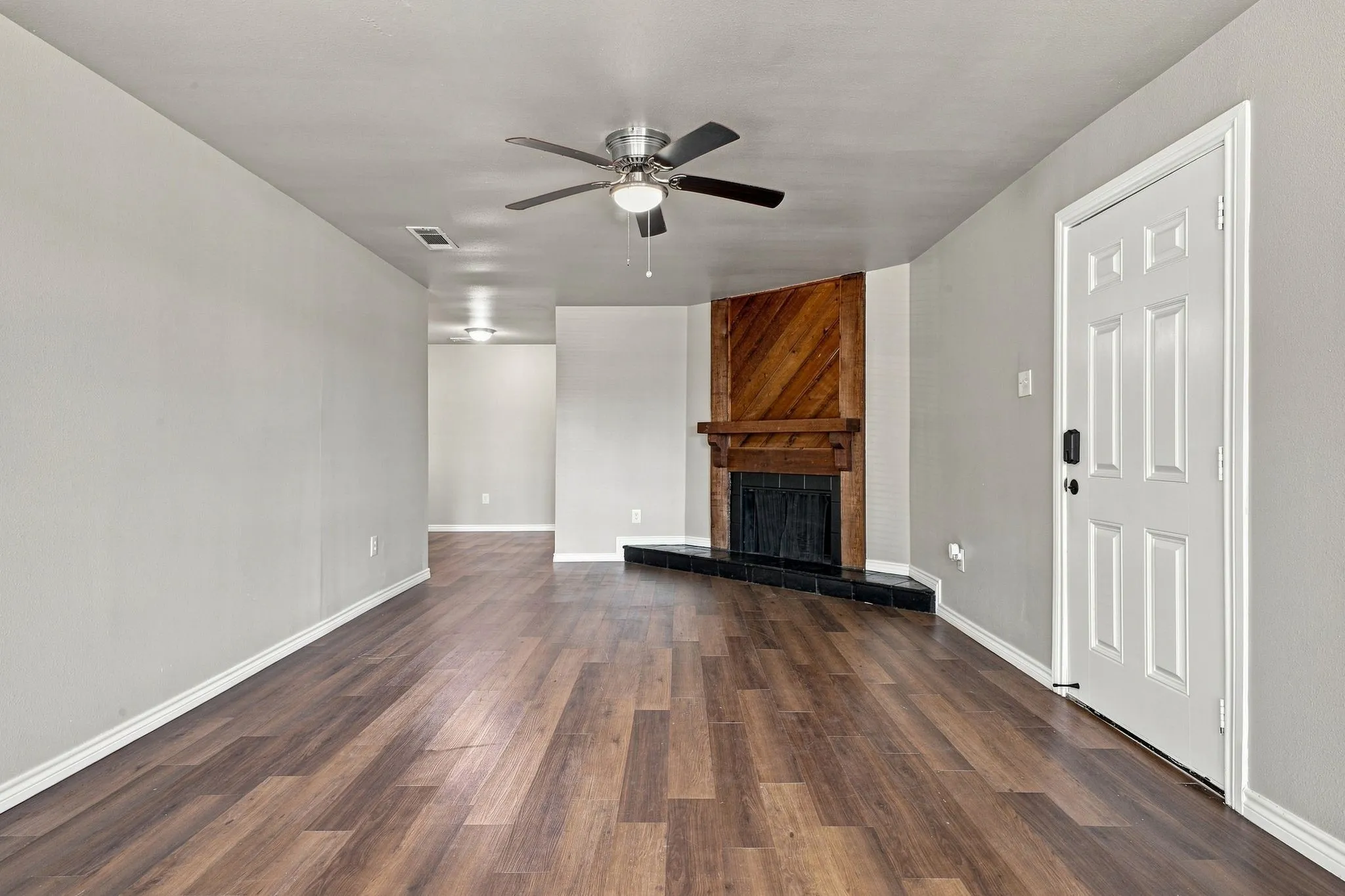 Spacious living area featuring wood-finish flooring, a corner fireplace with a wood-paneled surround and black hearth, and a ceiling fan with integrated lighting