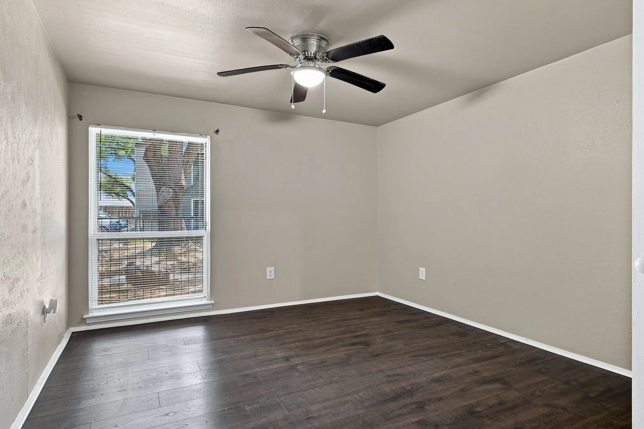Wood-finish flooring, neutral wall tones, ceiling fan with integrated lighting, and a single pane window