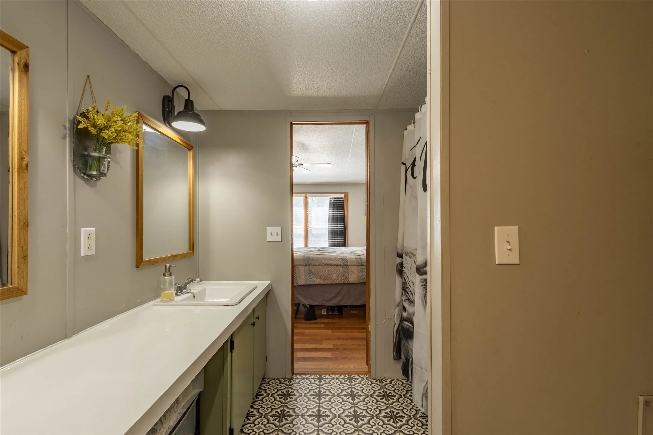 Extended vanity with an integrated sink and ample counter space, green wood-finish cabinetry, two framed mirrors, wall-mounted light fixtures, and patterned floor tile