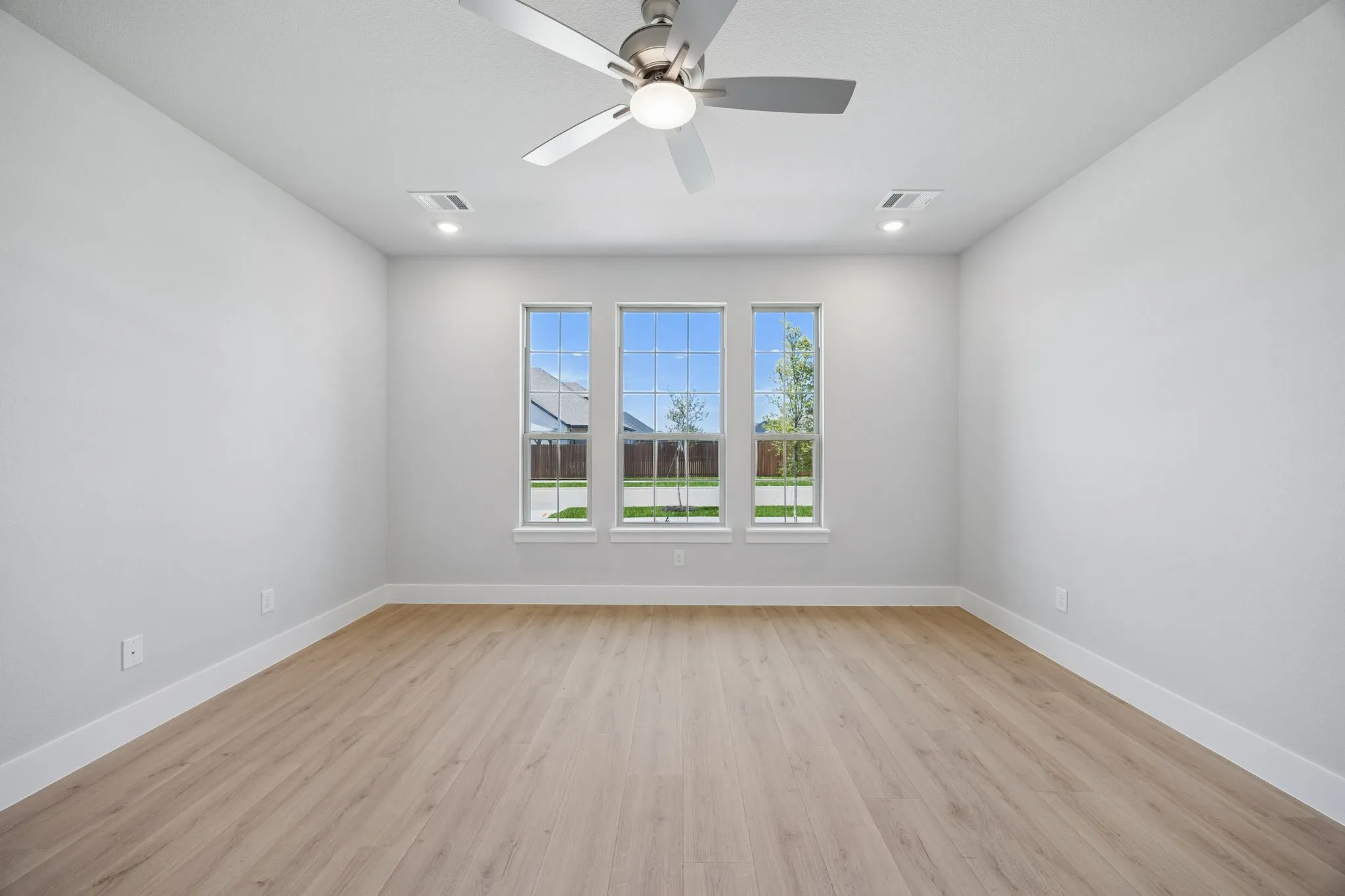 Bright, open room featuring expansive triple-pane windows with natural light, light wood-finish flooring throughout, ceiling fan with integrated lighting, and clean white walls with neutral finishes