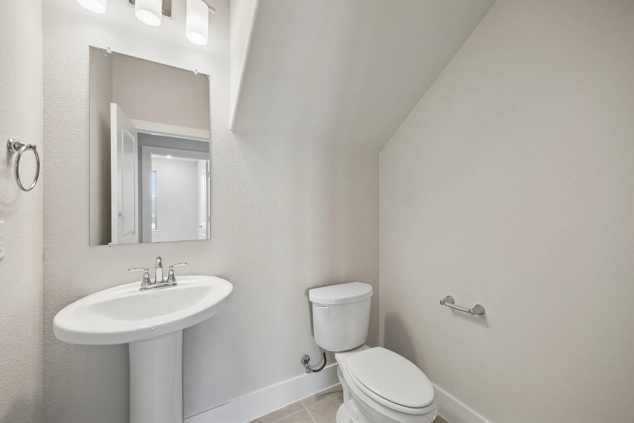 Pedestal sink with chrome fixtures, wall-mounted mirror, ceramic toilet, three-light vanity fixture, and light-toned floor tiling