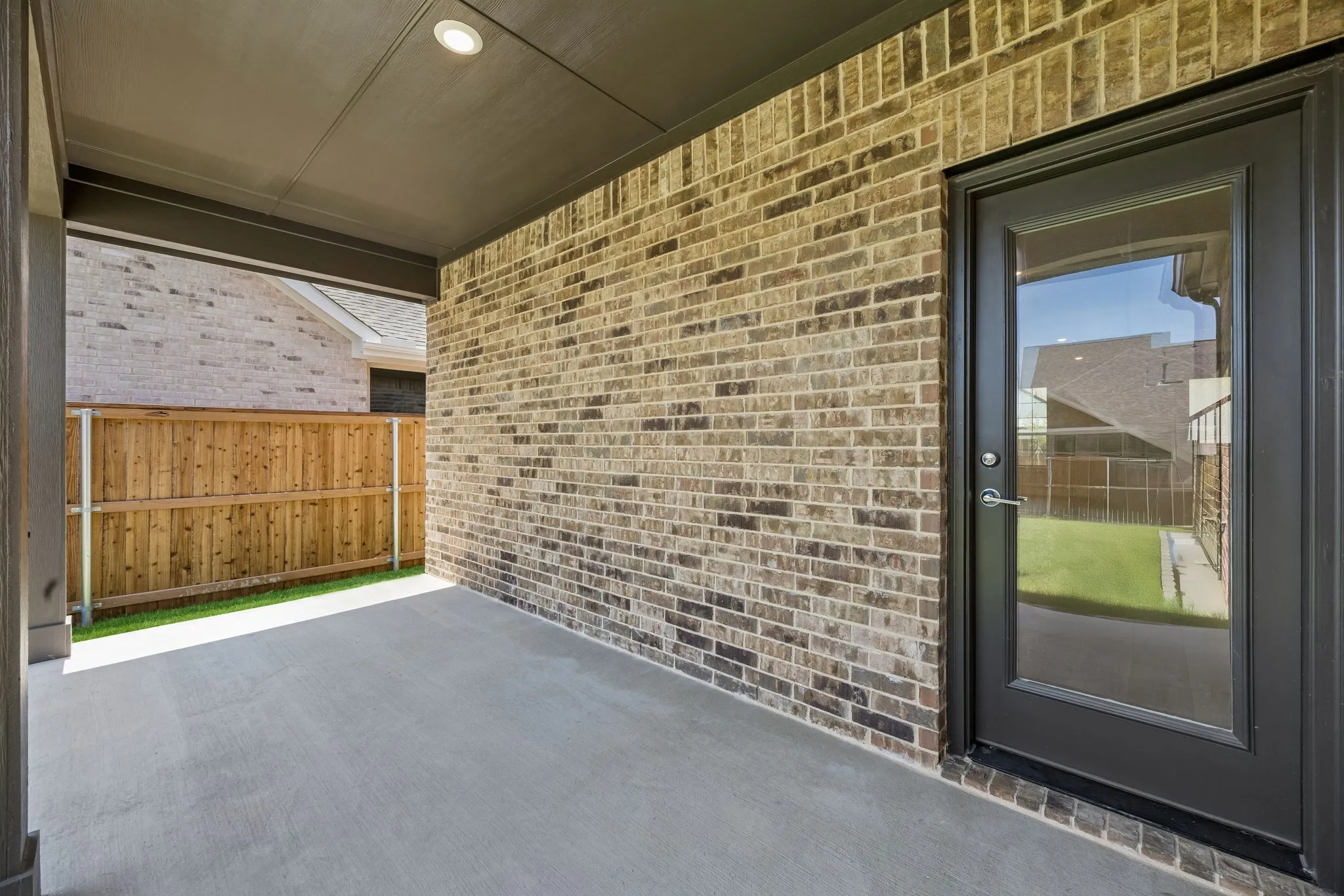 Covered patio featuring a recessed ceiling light, brick exterior wall, full-light door with dark trim, and a wood privacy fence