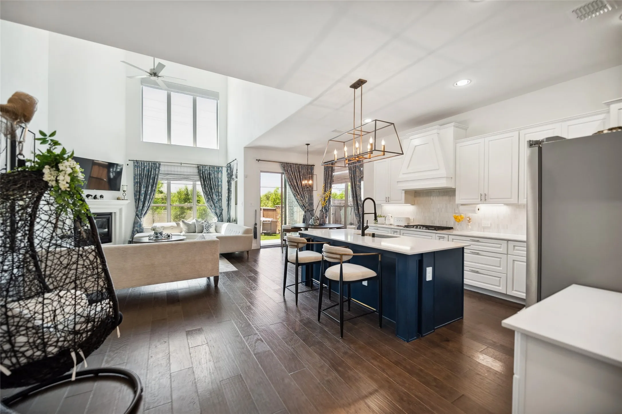 Wood-finish flooring throughout, kitchen island with seating, pendant lighting, white cabinetry, and a double-height living area with a fireplace