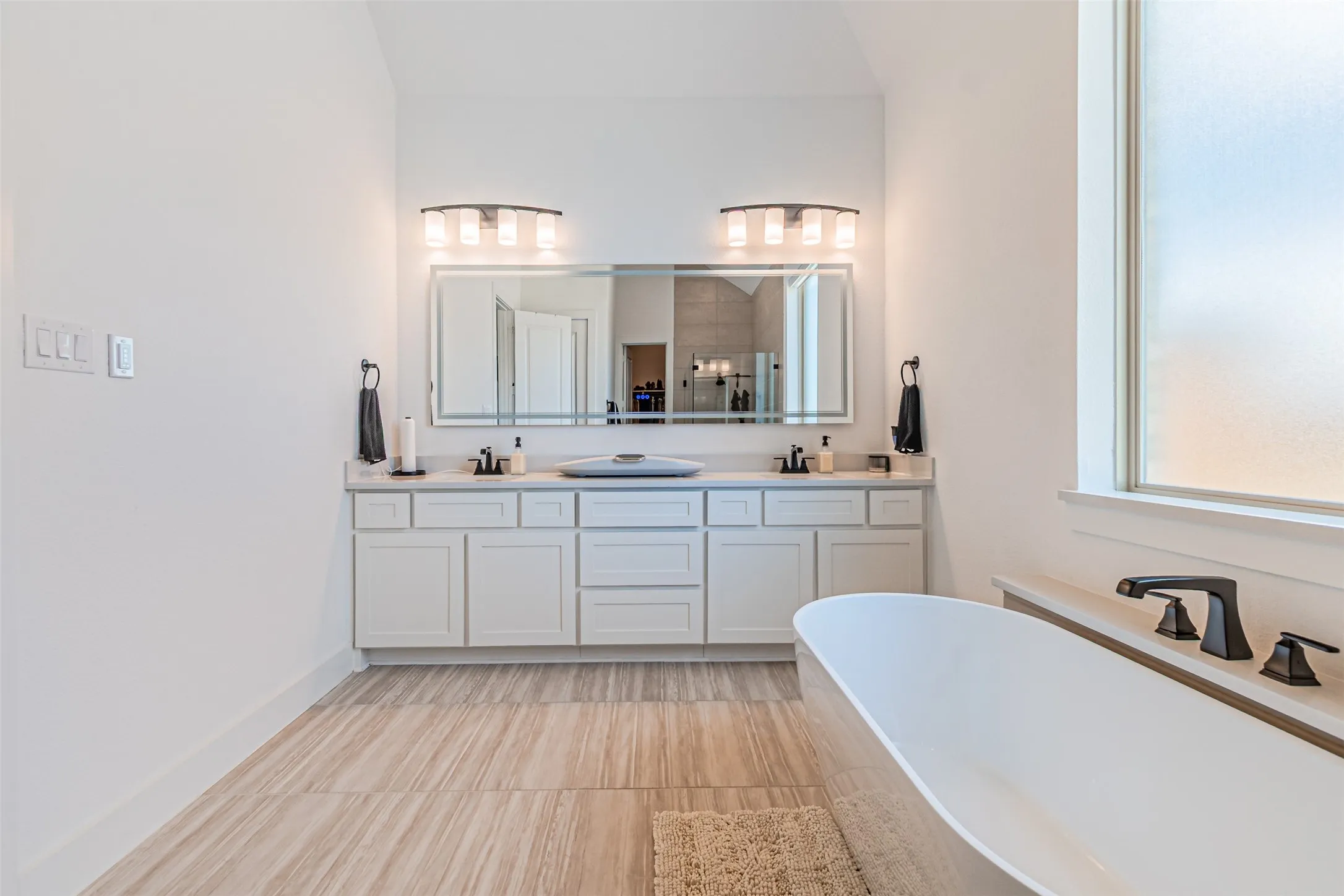 Dual vanity with white cabinetry and integrated sinks, matte black fixtures, freestanding soaking tub, and large frosted window