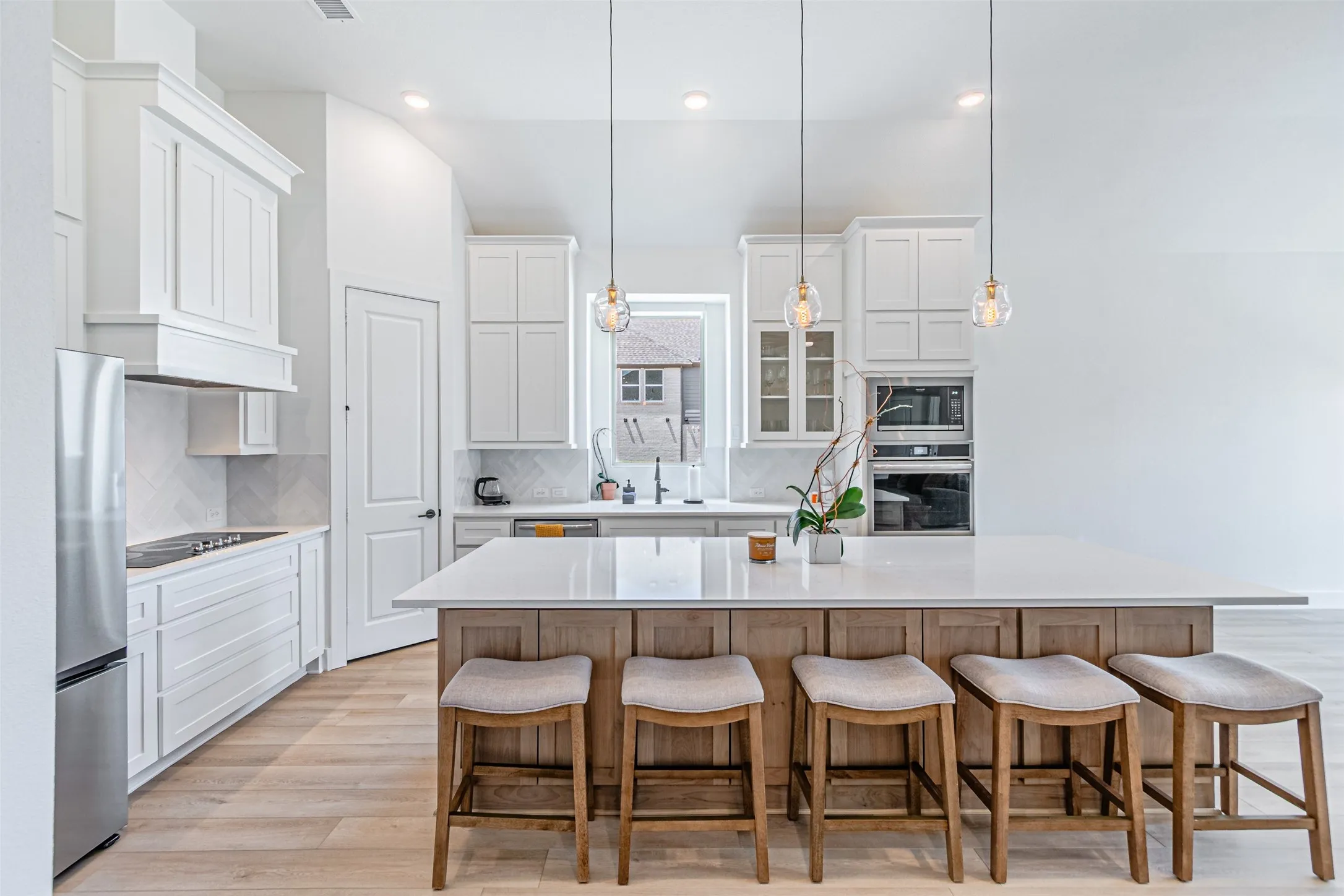 Gourmet kitchen featuring a large central island with seating, built-in stainless steel appliances, white cabinetry, a herringbone tile backsplash, and wood-finish flooring