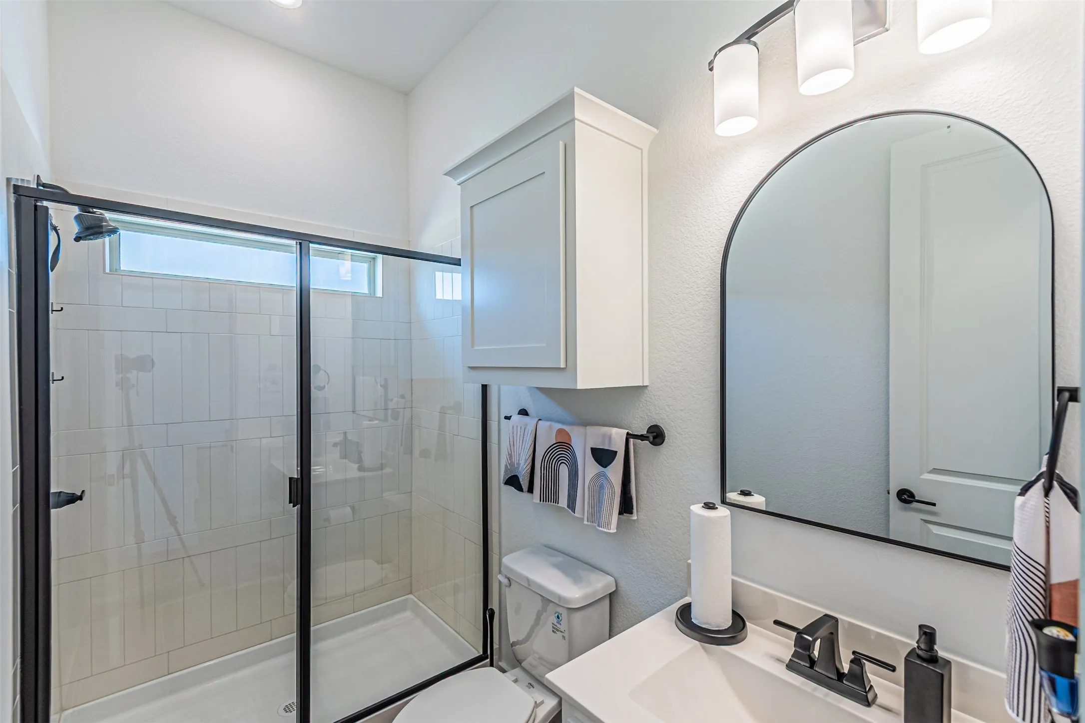 Walk-in shower with clear glass enclosure and subway tile surround, white vanity with integrated sink and chrome faucet, arched wall mirror, and built-in storage cabinet