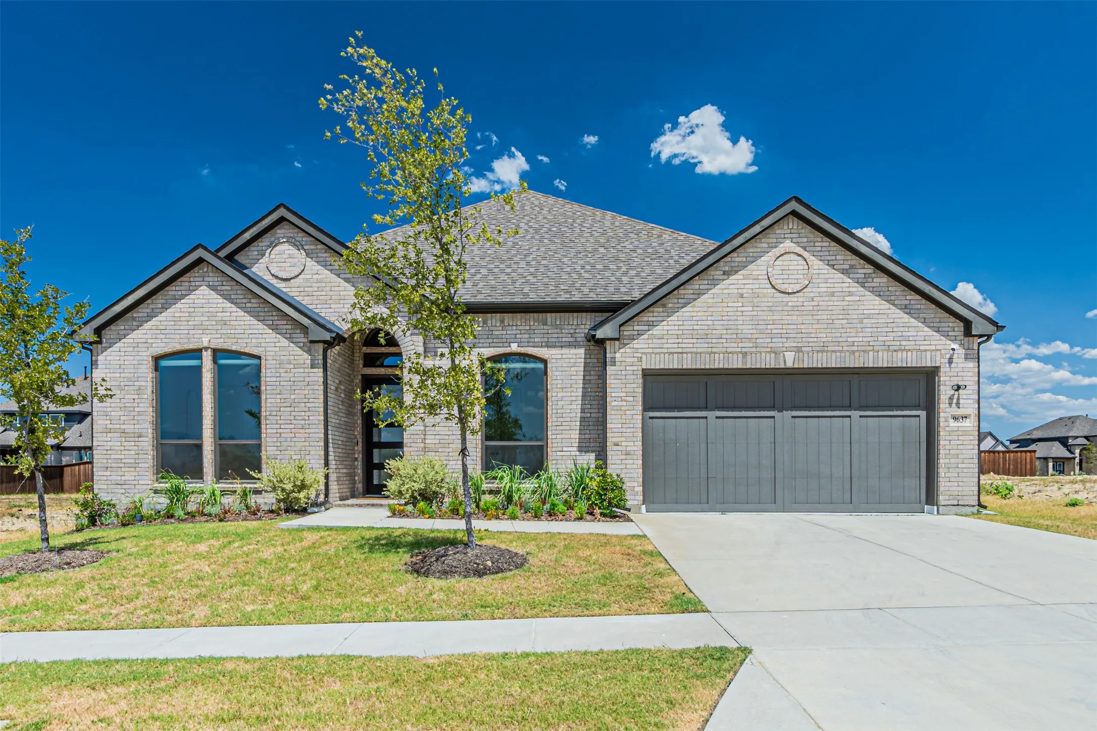 Light-colored brick exterior with dark trim, prominent gabled rooflines, arched entry, and a two-car garage with a panel-style door