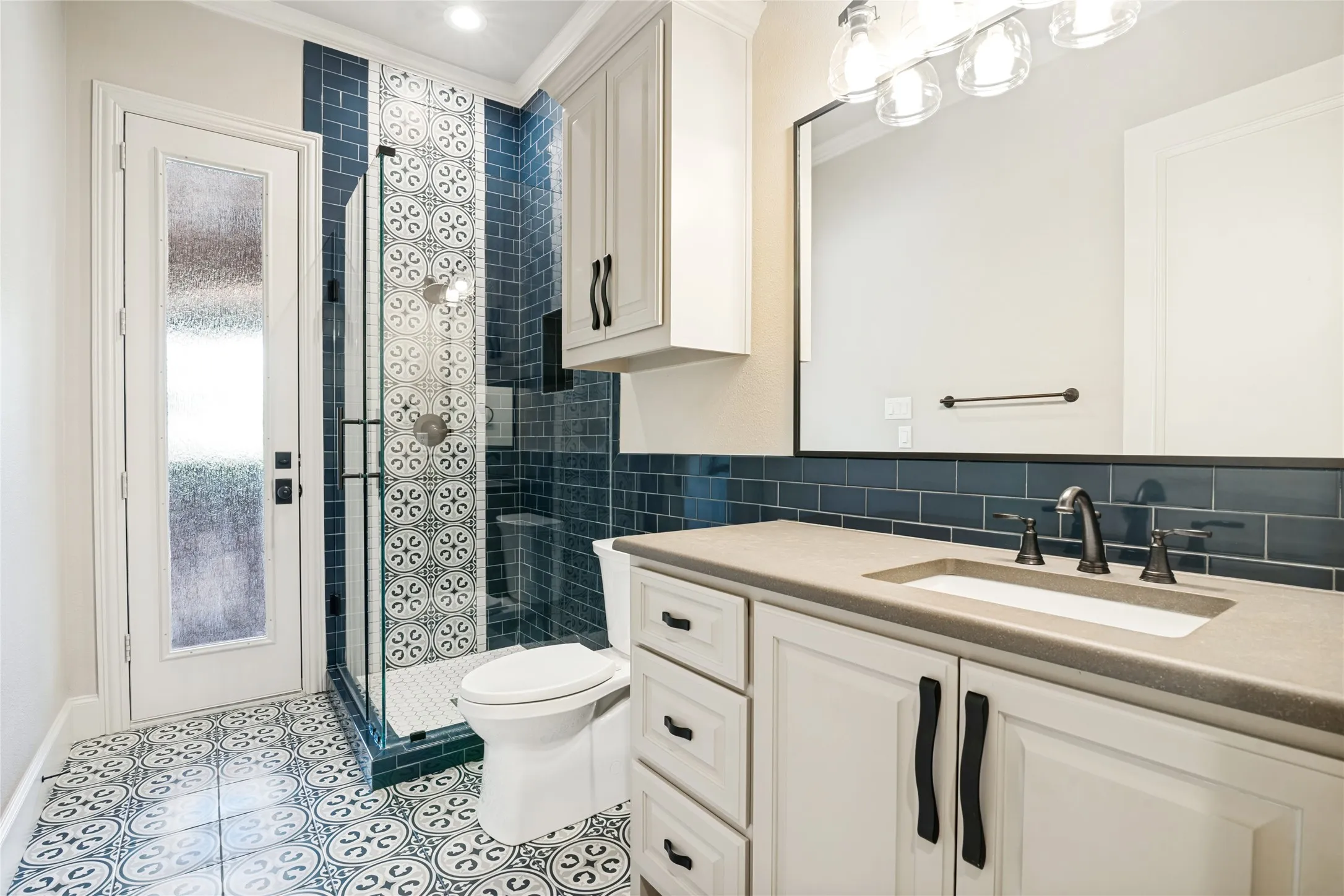 Patterned floor tiling, custom vanity with integrated sink, glass-enclosed shower with decorative tile, subway tile shower surround, and white cabinetry with dark hardware