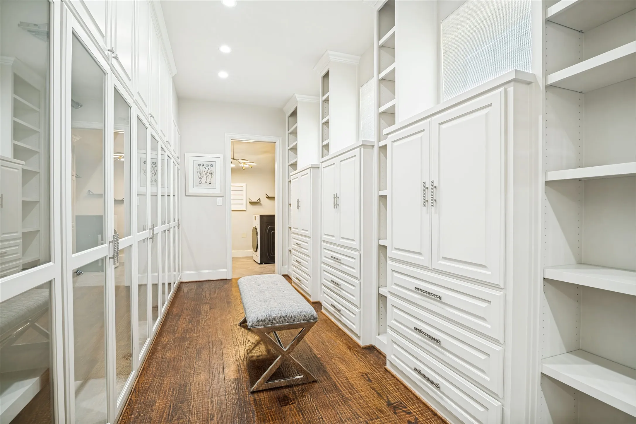 Luxurious walk-in closet featuring floor-to-ceiling white cabinetry with glass-front doors, open shelving, and wood-finish flooring