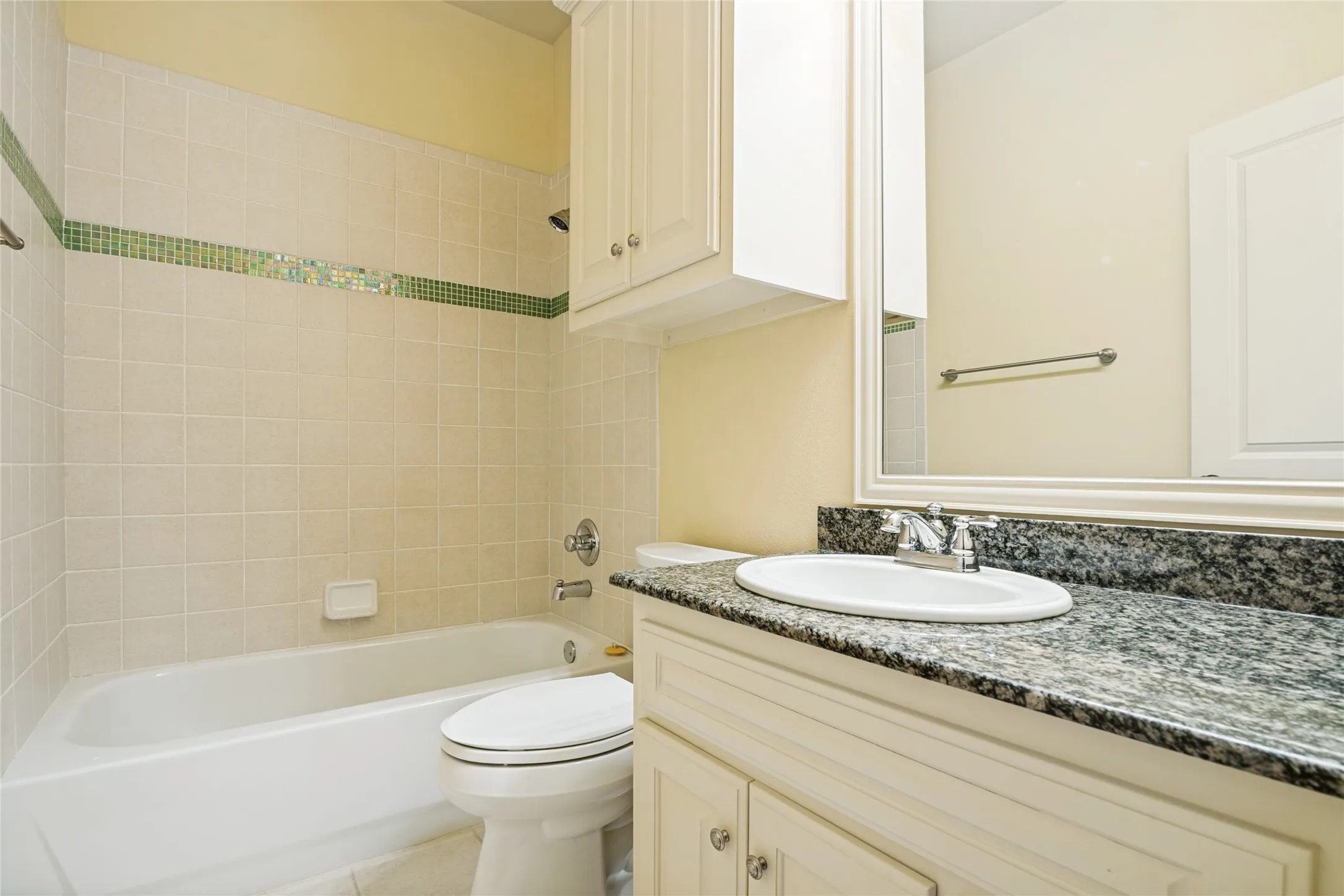 Bathroom featuring a white ceramic vessel sink with dark speckled stone countertop, white cabinetry with hardware accents, a large framed mirror, white subway tile walls with green tile accent band, and a white porcelain toilet and soaking tub