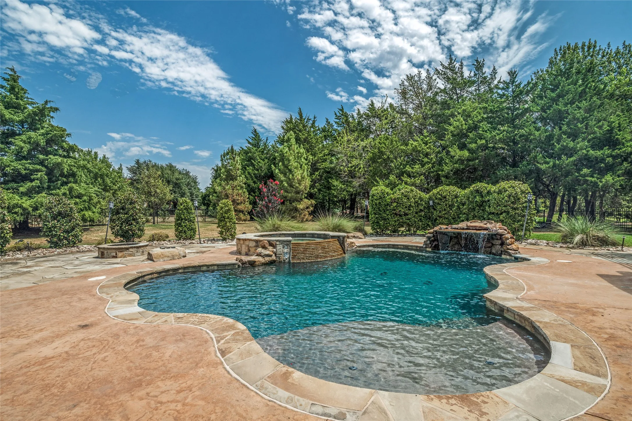 Freeform swimming pool with integrated spa and rock waterfall feature, surrounded by stamped concrete decking with stone coping
