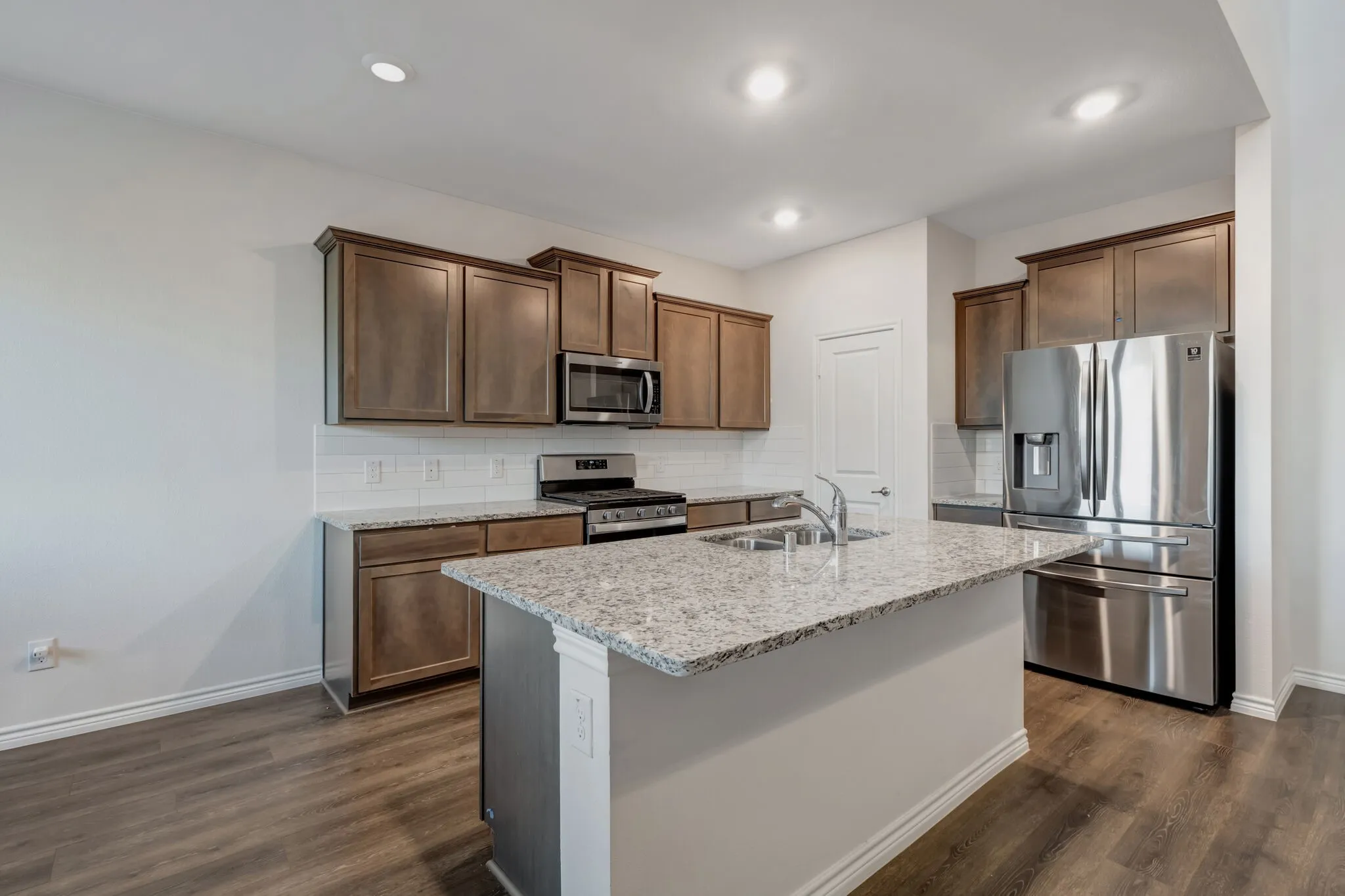 Kitchen featuring wood-finish flooring, wood cabinetry, granite countertops, a white subway tile backsplash, and stainless steel appliances