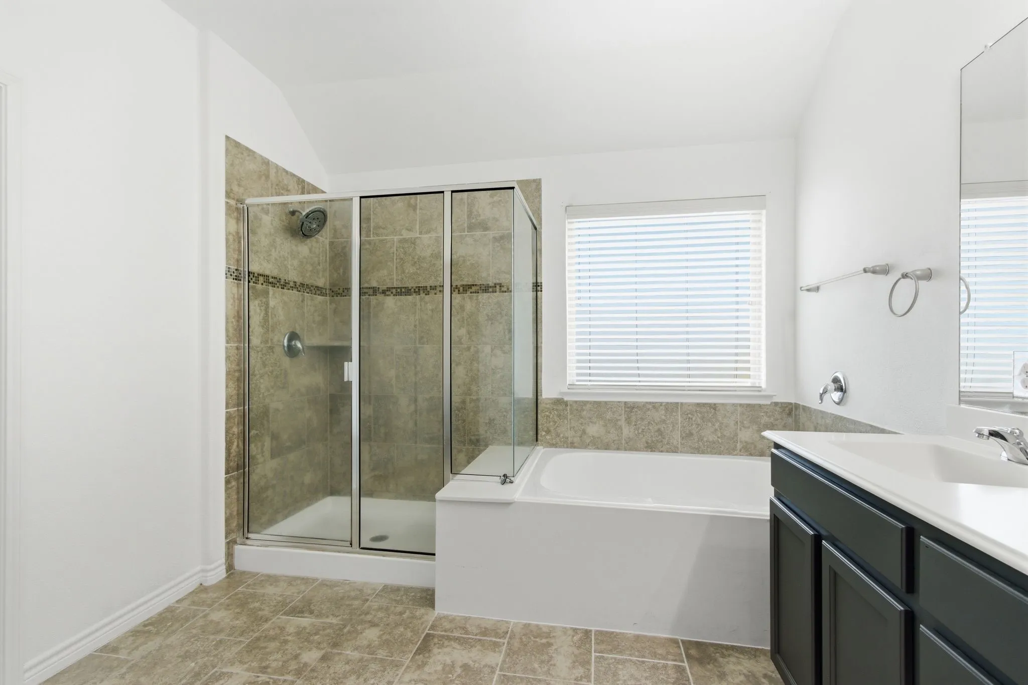 Tiled flooring, glass-enclosed shower with tiled surround, built-in soaking tub with tiled surround, single vanity with integrated sink, and a window with blinds