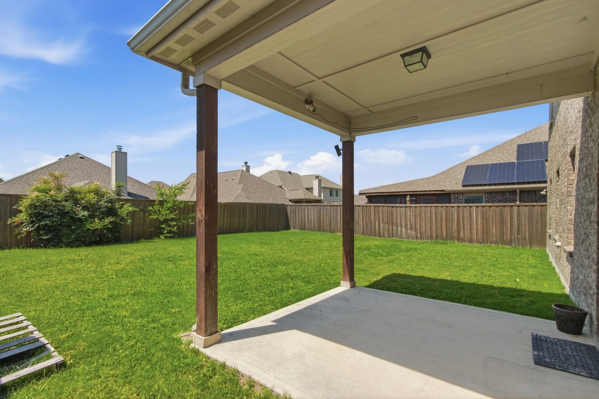 Covered concrete patio with overhead lighting, brick exterior, and a fully fenced backyard featuring a manicured lawn