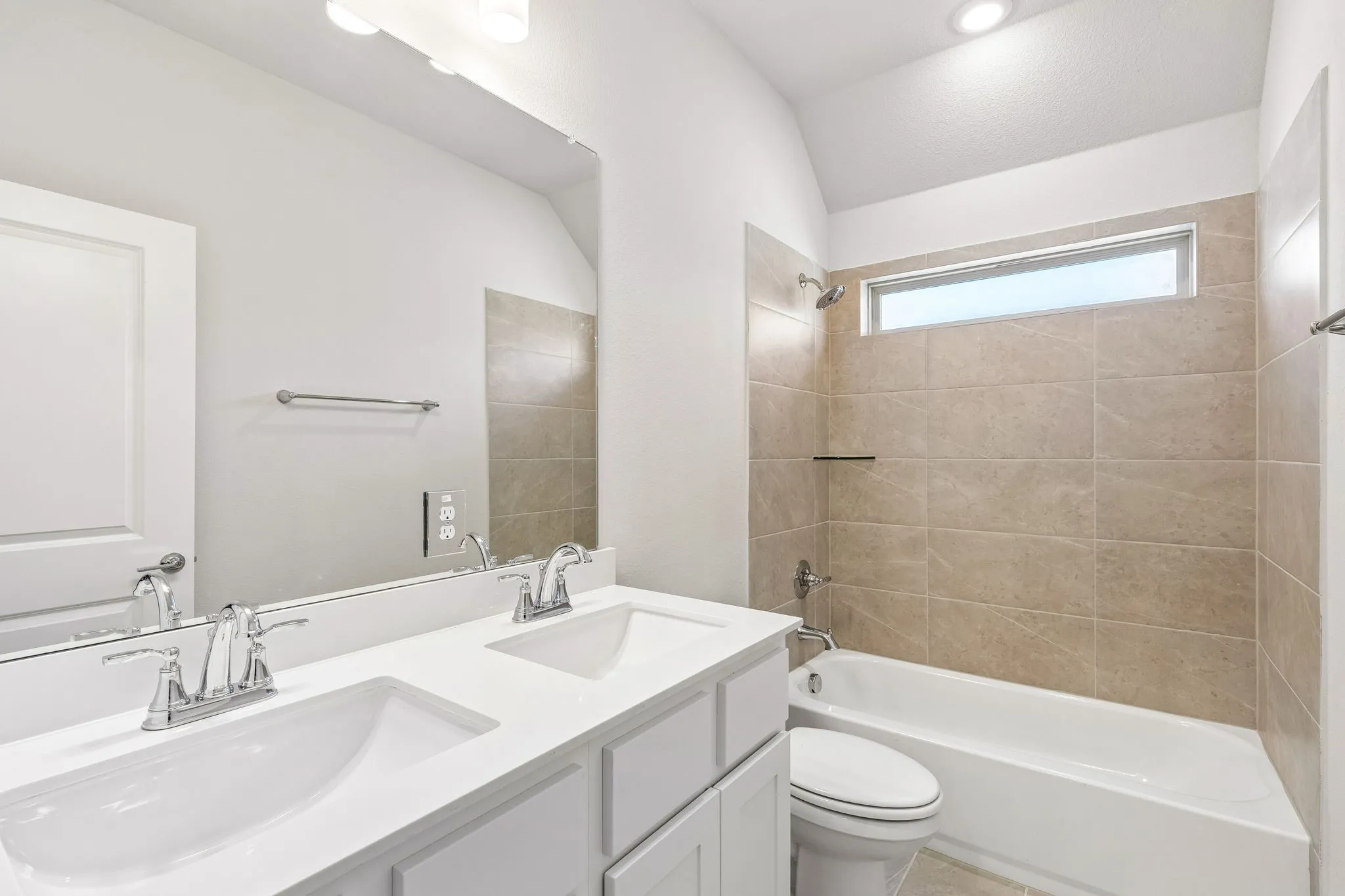 Dual vanity with chrome fixtures and white countertops, large mirror, tile-surround bathtub with shower, and a horizontal window
